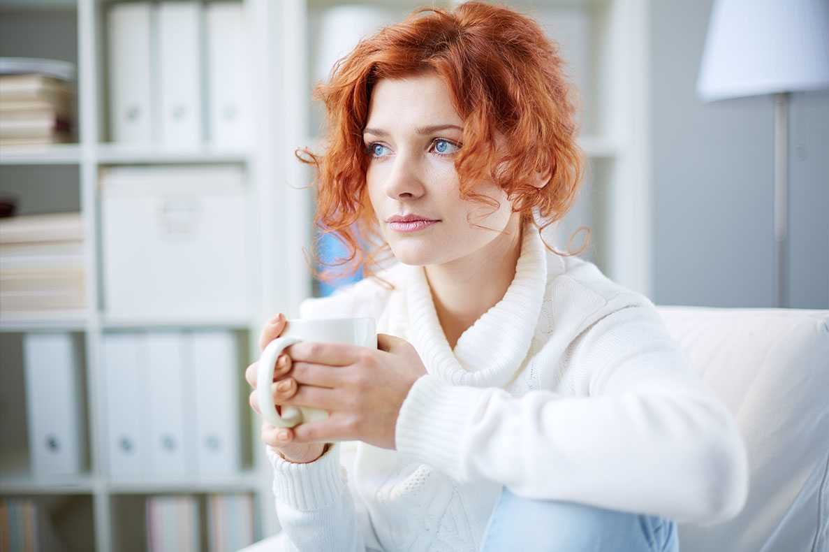 Mulher ruiva de olhos azuis sentada num sofá com uma caneca na mão e um ar preocupado